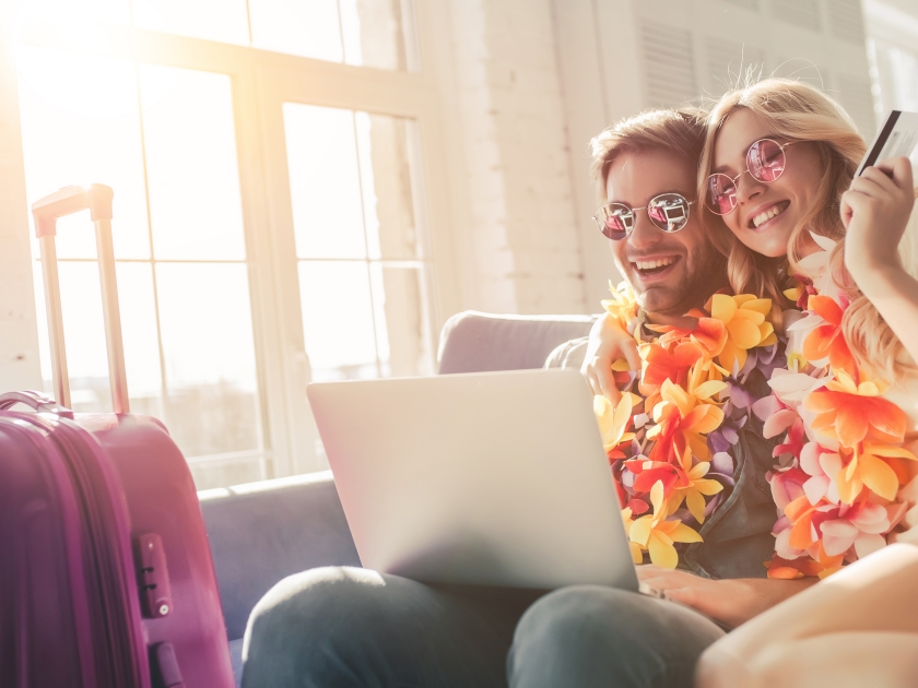 Young romantic couple wearing Hawaii accessories and sunglasses is preparing for travel at home. Sitting on sofa with laptop and credit card in hands while suitcases are standing nearby