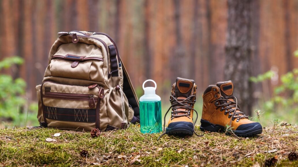 Hiking and camping equipment in forest. Backpack, water bottle and leather ankle boots. Panoramic view with copy space