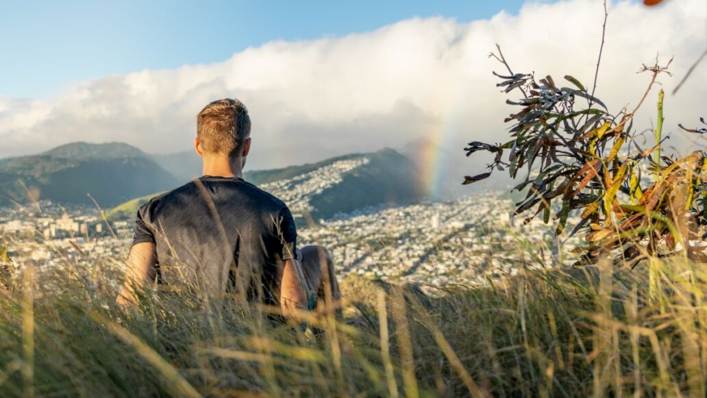 Young male hiker sitting on the summit of Diamond Head Crater in Honolulu on the Island of Oahu, Hawaii shortly before sunset, looking at a rainbow over the city and mountain range behind Honolulu.