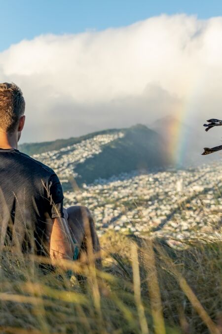 Young male hiker sitting on the summit of Diamond Head Crater in Honolulu on the Island of Oahu, Hawaii shortly before sunset, looking at a rainbow over the city and mountain range behind Honolulu.