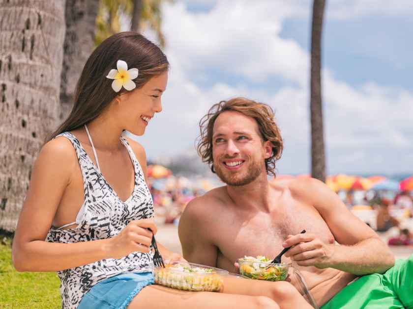 Eating salad couple having healthy snack takeout picnic lunch at beach after fun summer lifestyle. Young Asian woman, Caucasian man.