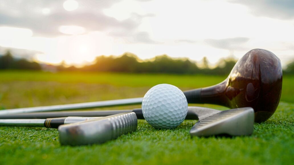 Close up old golf club and golf ball on green grass with sunset background. Golf ball and wooden driver on green grass background.