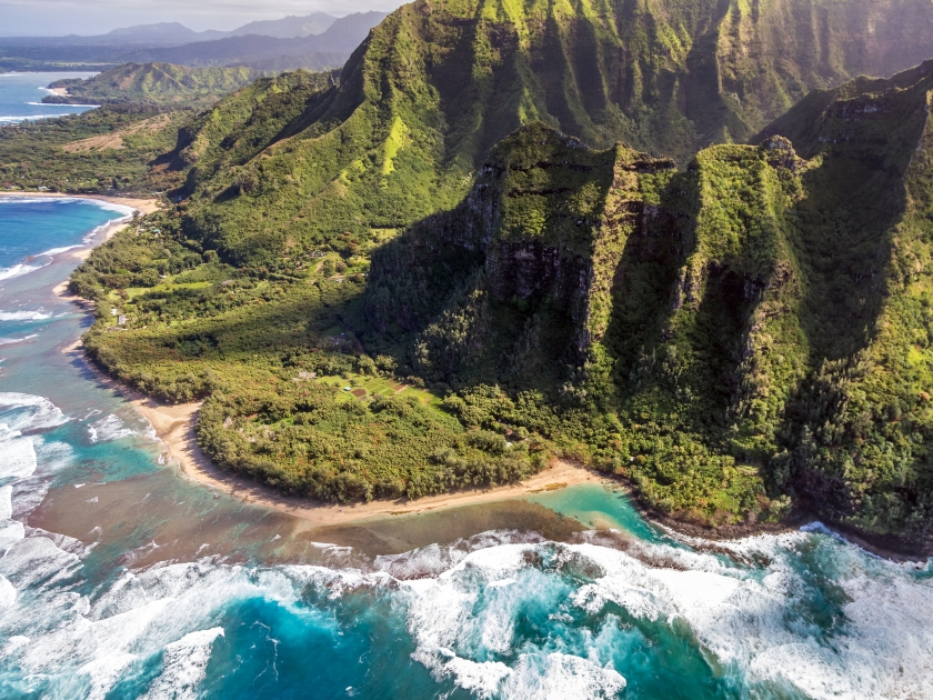 Aerial view of Kee Beach with tall, lush mountains of the Na pali coast and big waves crashing on the reef while on a doors off helicopter tour of Kauai, Hawaii.