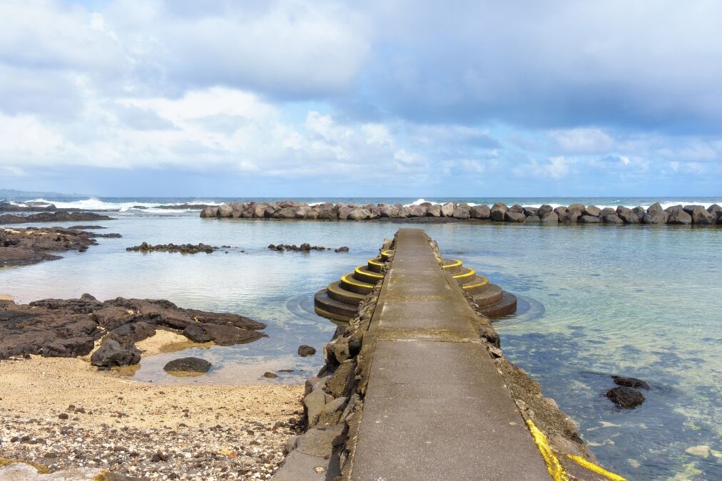 Beautiful Hawaii beach with a sandy shoal and a stone pier with steps for easy access to the water, separated from the ocean by a massive stone barrier.