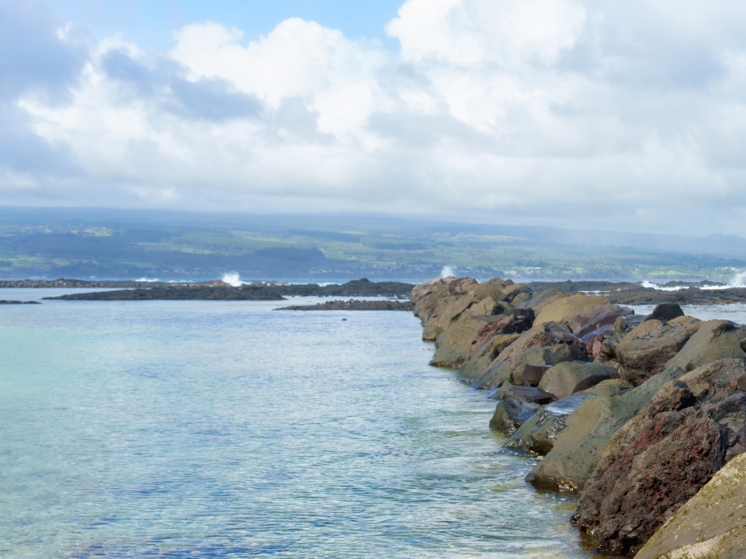 Picturesque scene of a Hawaiian beach with large rocks dividing the shoreline from the ocean, framed by an endless ocean horizon and rolling clouds above.