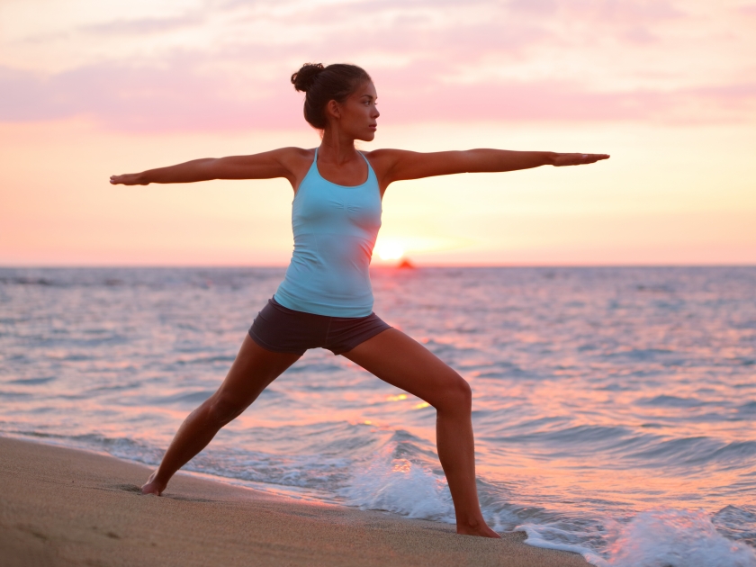 Yoga woman in zen meditating in warrior pose relaxing outside by beach at sunrise or sunset. Female yoga instructor working out training in serene ocean landscape. Big Island, Hawaii, USA.