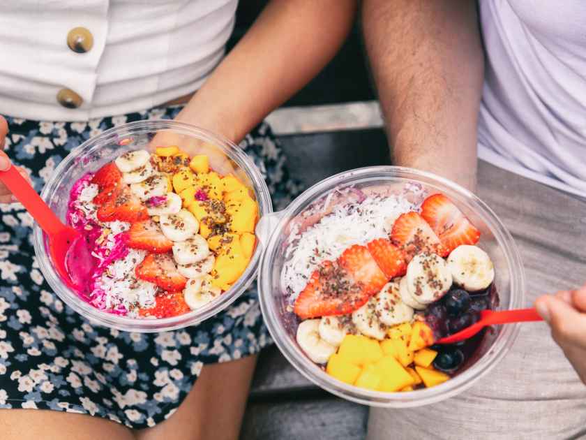 Acai bowl and pitaya dragonfruit smoothie healthy breakfast bowls young friends eating together. Couple man and woman eating sitting outside in park for lunch break. Closeup on food.