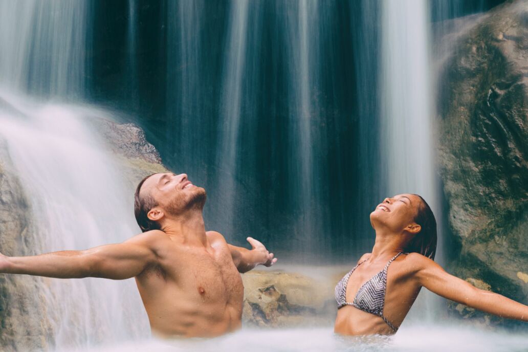 Nature,Couple,Relaxing,Under,Waterfall,With,Open,Arms,In,Freedom