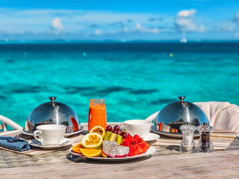 Vacation breakfast at luxury hotel room ocean view. Romantic honeymoon travel holiday in Maldives or Tahiti. Presentation of two plates, fruits, coffee, juice.