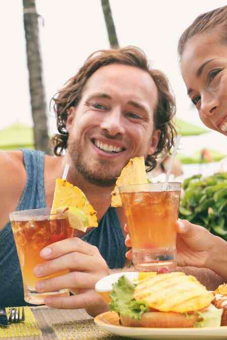 Selfie photo couple taking picture with phone at Hawaii beach bar on summer vacation. Happy Asian woman and man toasting mai tai drinks at bar having fun. Friends on travel holidays.