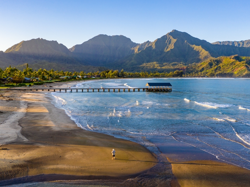 Aerial image at sunrise off the coast over Hanalei Bay and pier on Hawaiian island of Kauai with a man standing alone on the beach