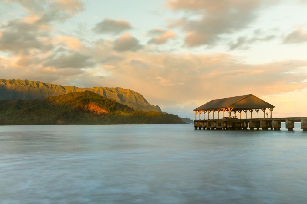 Rising sun illuminates the peaks of Na Pali mountains over the calm bay and Hanalei Pier in long exposure photo