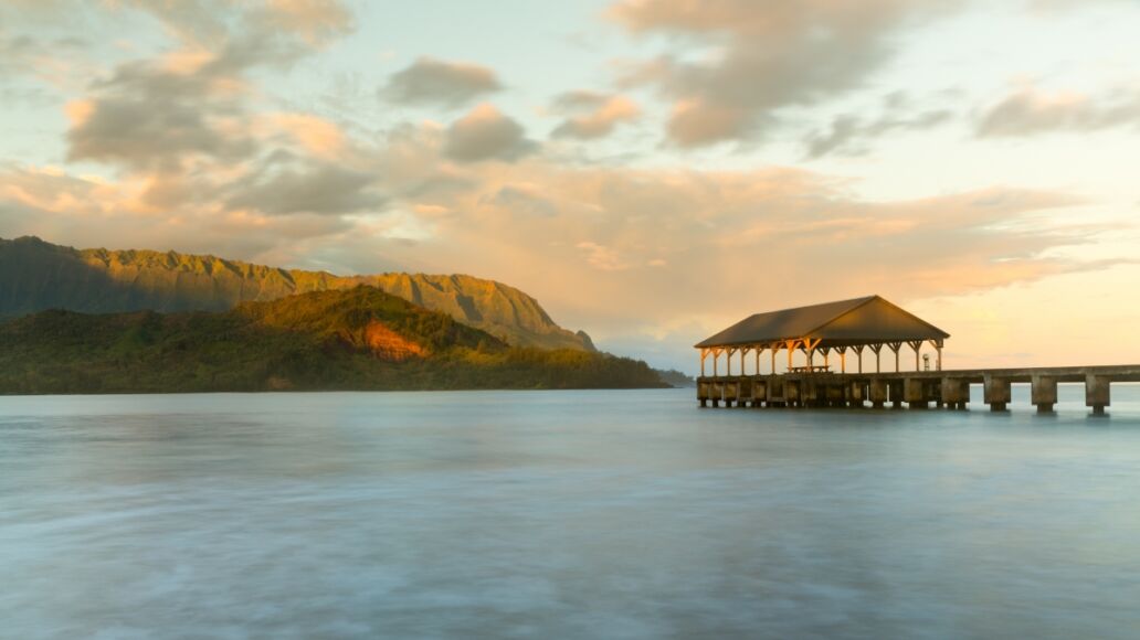 Rising sun illuminates the peaks of Na Pali mountains over the calm bay and Hanalei Pier in long exposure photo