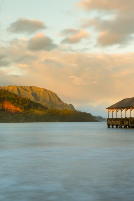 Rising sun illuminates the peaks of Na Pali mountains over the calm bay and Hanalei Pier in long exposure photo