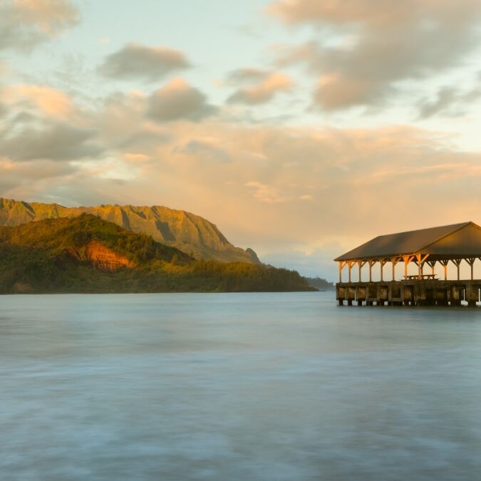 Rising sun illuminates the peaks of Na Pali mountains over the calm bay and Hanalei Pier in long exposure photo