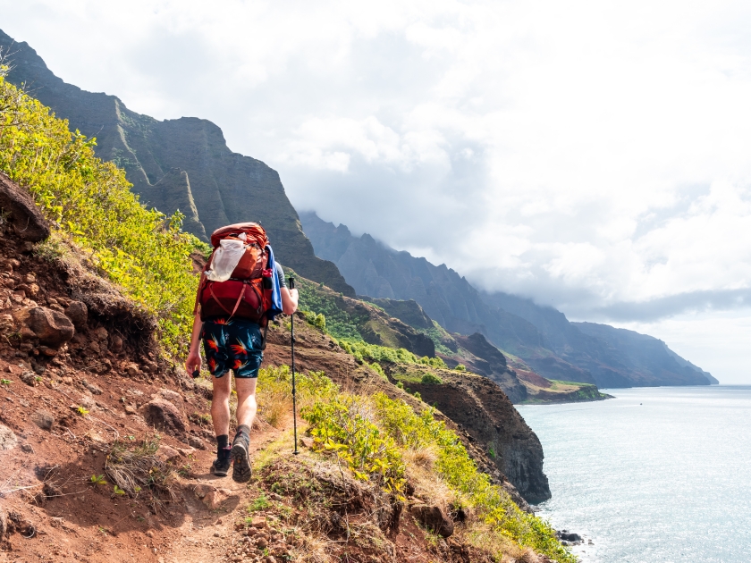 Hiker on a treacherous coastal trail with valleys and cliffs on one side and the ocean on the other. Shot on the Kalalau Trail in the Na Pali Coast State Park on the island of Kauai, Hawaii USA.