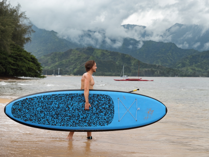 Stand up paddleboard fit man carrying paddle board going on ocean swim watersport activity at Puu Poa beach, Hanalei Bay, Kauai, Hawaii, USA. Hawaii travel sport athlete.