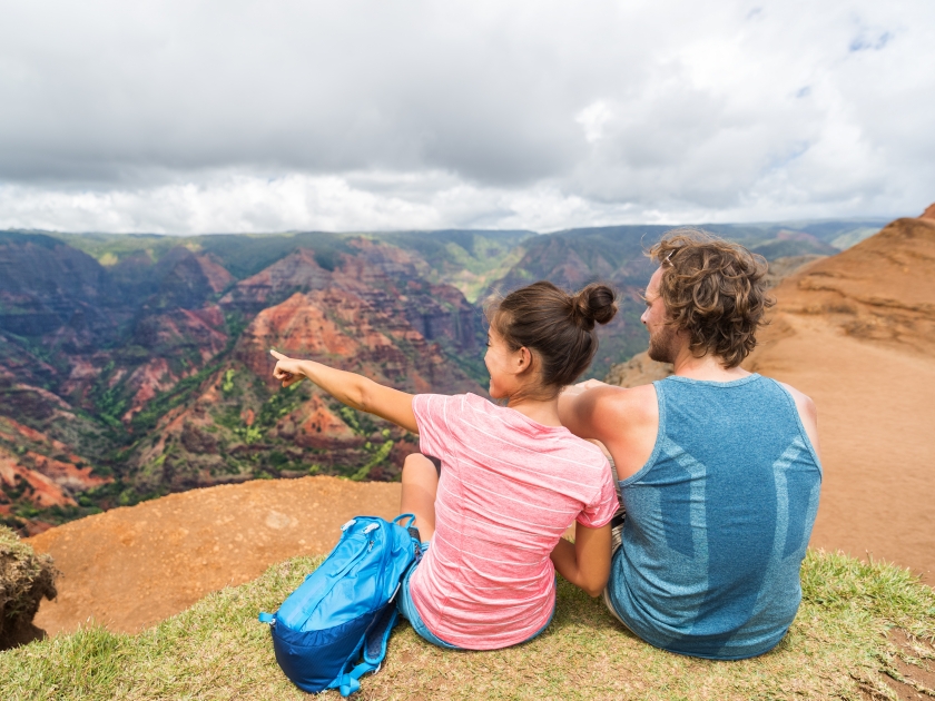 People hiking in Hawaii hikers pointing at Kauai. happy hiker couple healthy lifestyle outdoors looking at Waimea canyon view. Young couple resting sitting in nature in Kauai, Hawaii, USA.