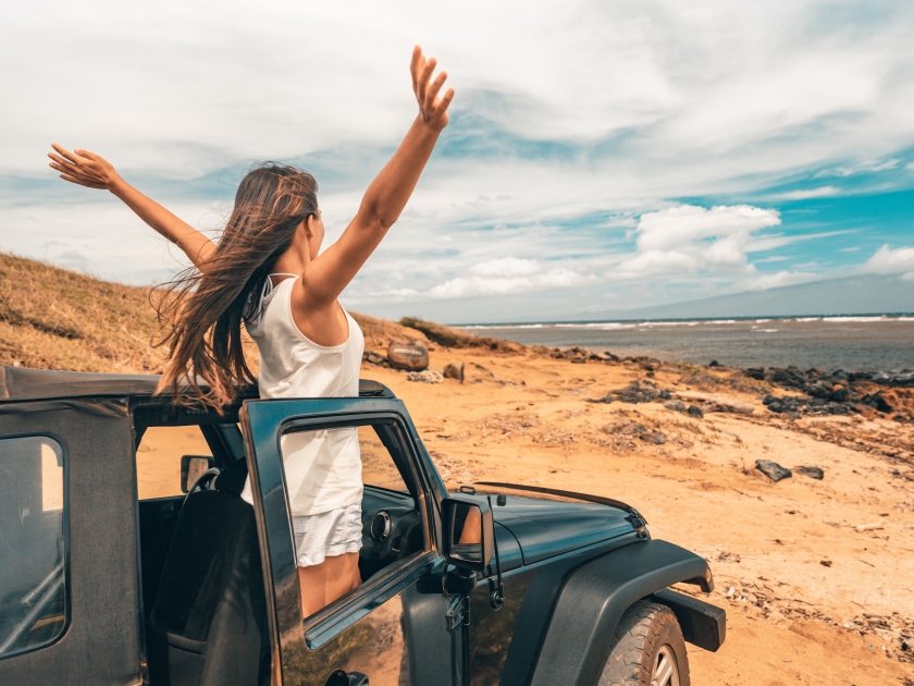 Car road trip travel fun happy woman tourist with open arms at ocean view from sports utility car driving on beach. Summer vacation adventure girl from the back.