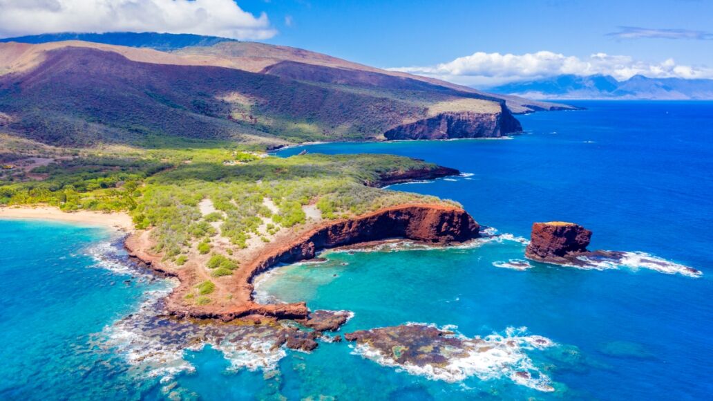 Aerial view of Lanai, Hawaii featuring Hulopo'e Bay and beach, Sweetheart Rock (Pu'u Pehe), Shark's Bay, and the mountains of Maui in the background.