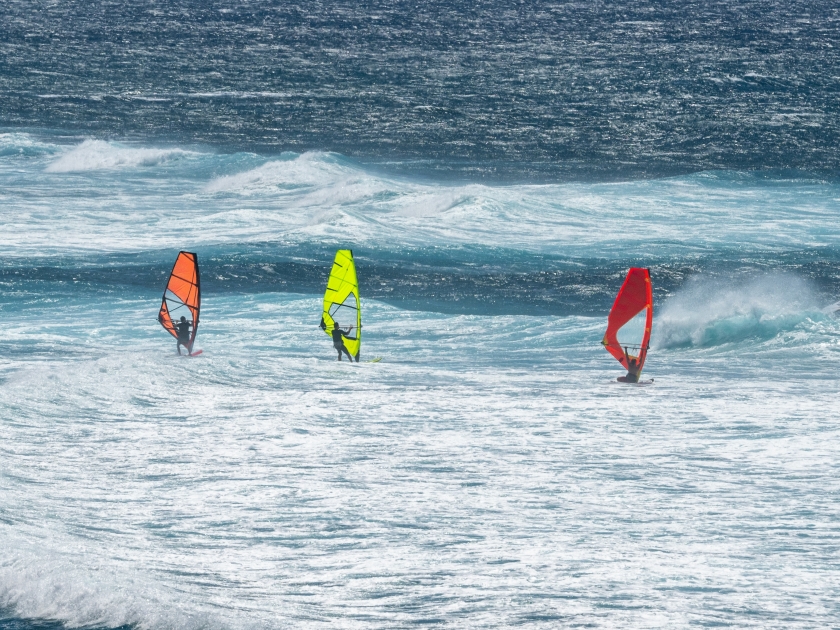 Three colorful windsurfers, red, orange, and yellow, racing in the wind on the Pacific Ocean off the coast of Maui at Hookipa Beach, Hawaii