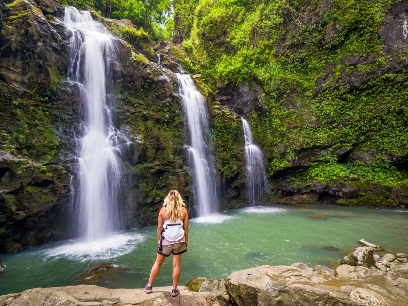 Blonde girl admires Three Bears Falls in Maui, Hawaii Hana Highway - Upper Waikani Falls. Road to Hana connects Kahului to the town of Hana Over 59 bridges, 620 curves, tropical rainforest.