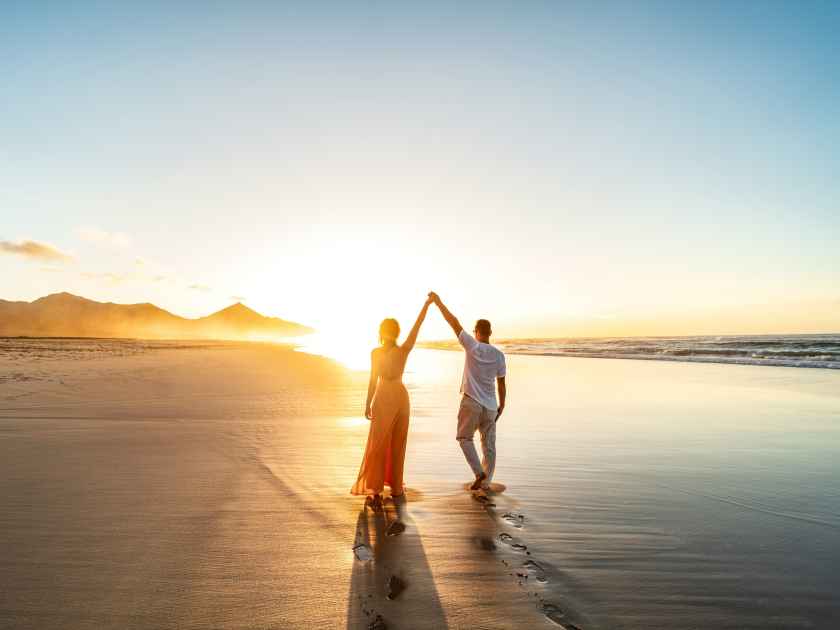 Lovely, romantic couple walking on sunset beach, enjoying evening light, relaxing on tropical summer vacation. Honeymoon. Love. Back view. Woman wearing orange maxi dress.