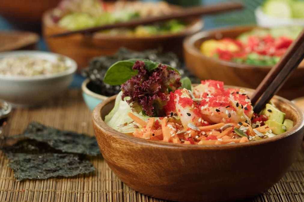 Traditional Hawaiian cuisine on table close-up. Cooked poke bowls served with sliced vegetables, fish and greenery, dried seaweed. Healthy vegetarian food. Asian vegan raw meal, chopsticks.