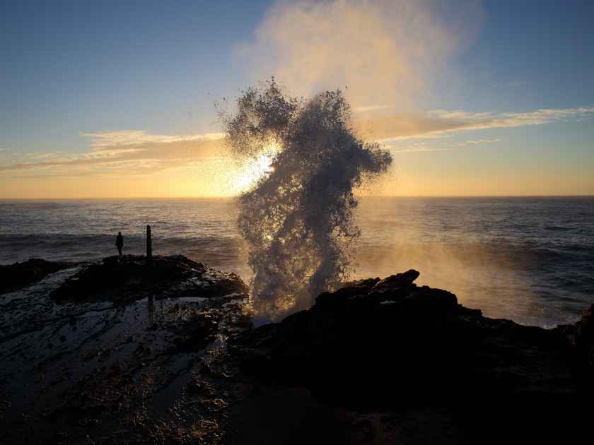 Water is shot up and out of a blowhole in the rocks in front of the early morning sunrise