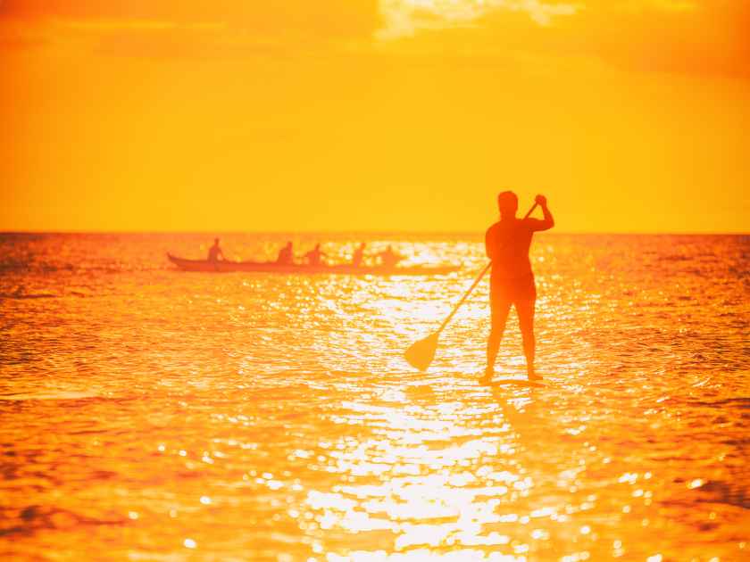 Hawaii ocean lifestyle - watersport activity on ocean - stand up paddleboard, people training on outrigger canoe . Active summer healthy living. Silhouette of standing person doing paddle board.