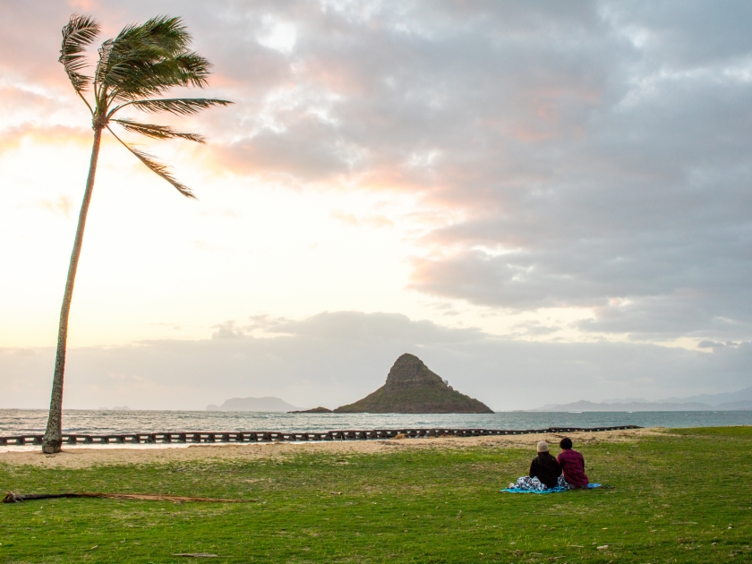 A couple sitting on grass watching overcast sunrise at beach park on a cool windy morning. Kualoa Regional Park, East Oahu, Hawaii, USA