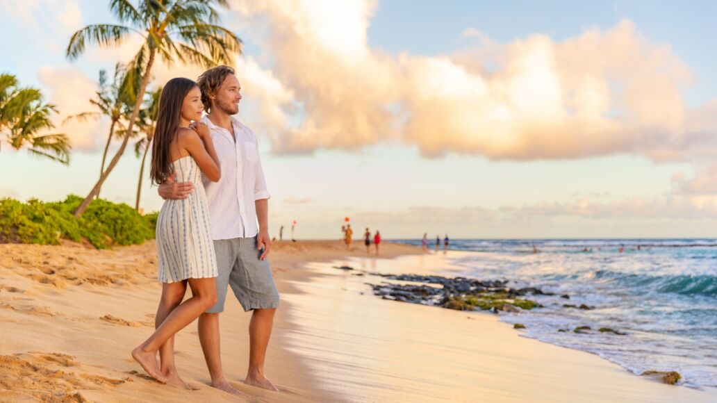 Couple on romantic sunset beach walk relaxing during Hawaii summer travel vacation on Kauai island, USA. Asian woman in white dress, Man in linen shirt.