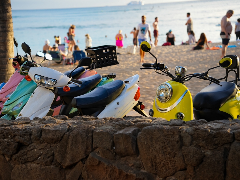 Multicolored scooters parked at Waikiki beach with beach goers, beach and ocean in background