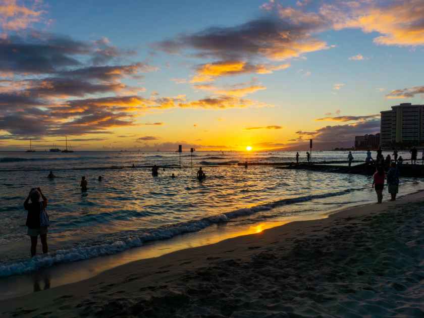 Golden sunset on horizon over Waikiki beach in Oahu, Hawaii, with silhouette of unidentifiable visitors swimming and on the beach.