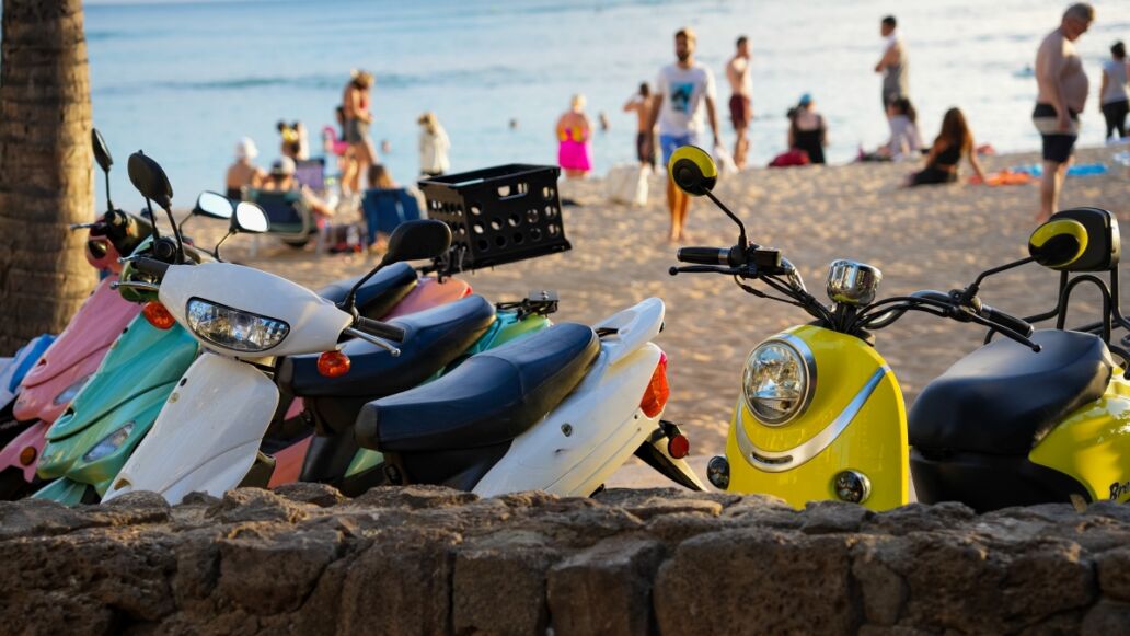 Multicolored scooters parked at Waikiki beach with beach goers, beach and ocean in background