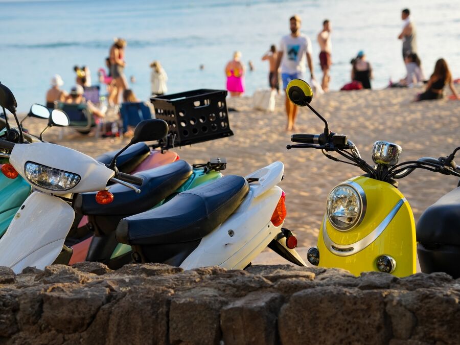 Multicolored scooters parked at Waikiki beach with beach goers, beach and ocean in background