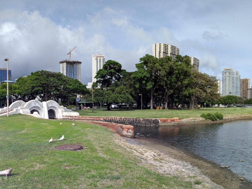 Waterway with bridge leads to pond in Ala Moana Beach Park surrounded by trees with Condominiums towers across the street on a nice day on Oahu, Hawaii.