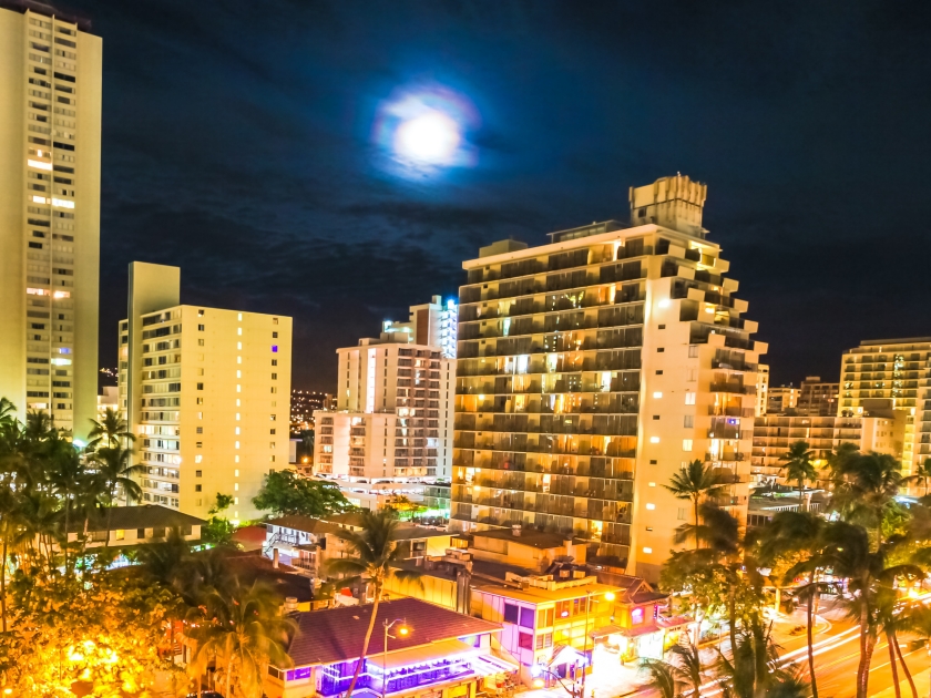 Moonlight aerial view on night traffic of Waikiki city in Oahu, Hawaii, United States. Moving people, car glowing trails in the street. City night lights of shops, shining full moon. Nightlife concept