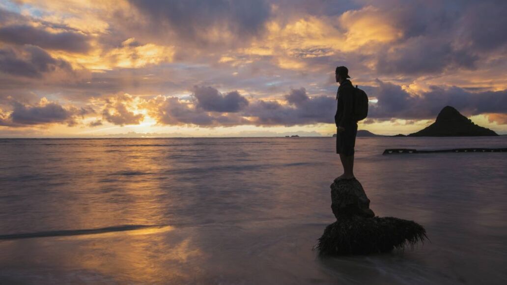 A person standing on a coconut tree stump in the shore line of Oahu's windward coast during a golden sunrise. The island known as China Man's Hat in the background.