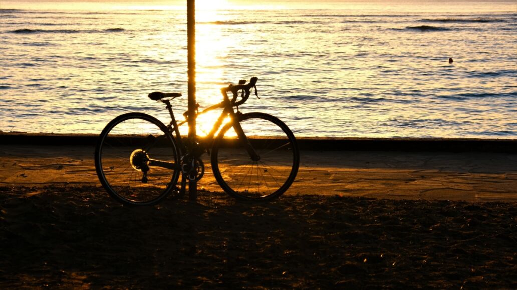 Golden sunset at Waikiki Beach with sailboats on the horizon. Silhouettes of hau tree, bicycle, palm trees and people. People silhouetted are watching sunset. Sky from light blue to yellow/orange.