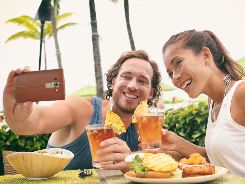 Selfie photo couple taking picture with phone at Hawaii beach bar on summer vacation. Happy Asian woman and man toasting mai tai drinks at bar having fun. Friends on travel holidays.