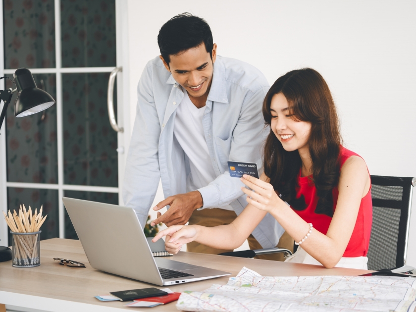 Young adult southeast asian couple using laptop and credit card buying airplane ticket for travel. People in love getting ready for holiday trip concept.