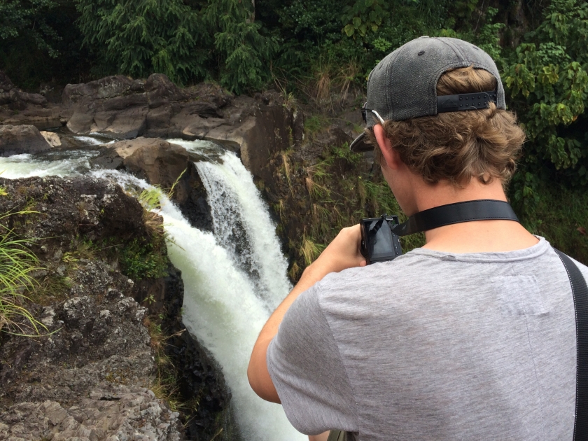 A young man with curly sandy blonde brown hair, a grey shirt and hat taking a photo on a camera of a beautiful natural waterfall off the edge of a rock cliff in The Big Island, Hawaii, Usa.